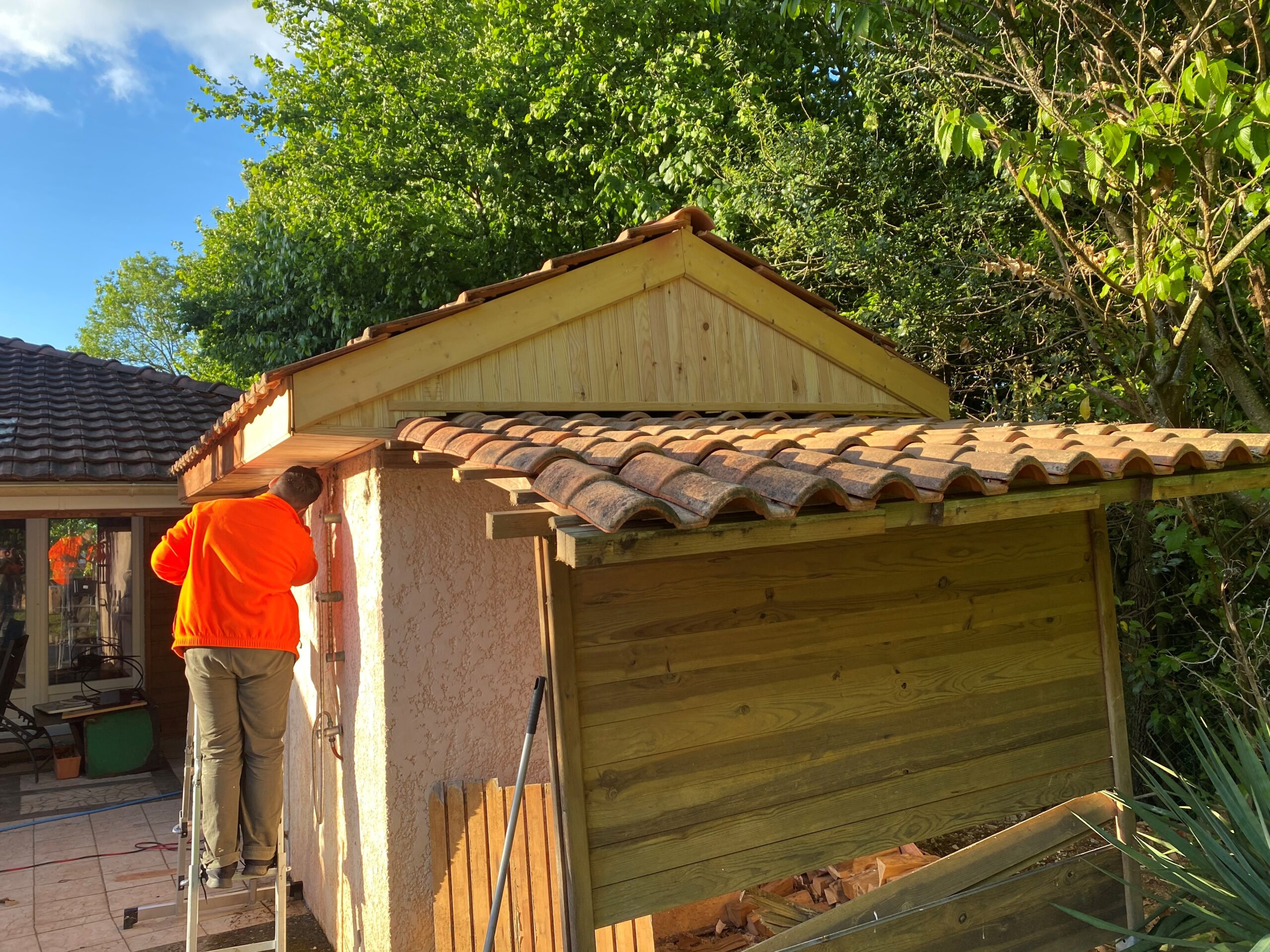 Ouvrier en haut d'un escabeau, vêtu d'un blouson orange, effectuant des travaux de toiture et de bardage en bois sur une extension extérieure.